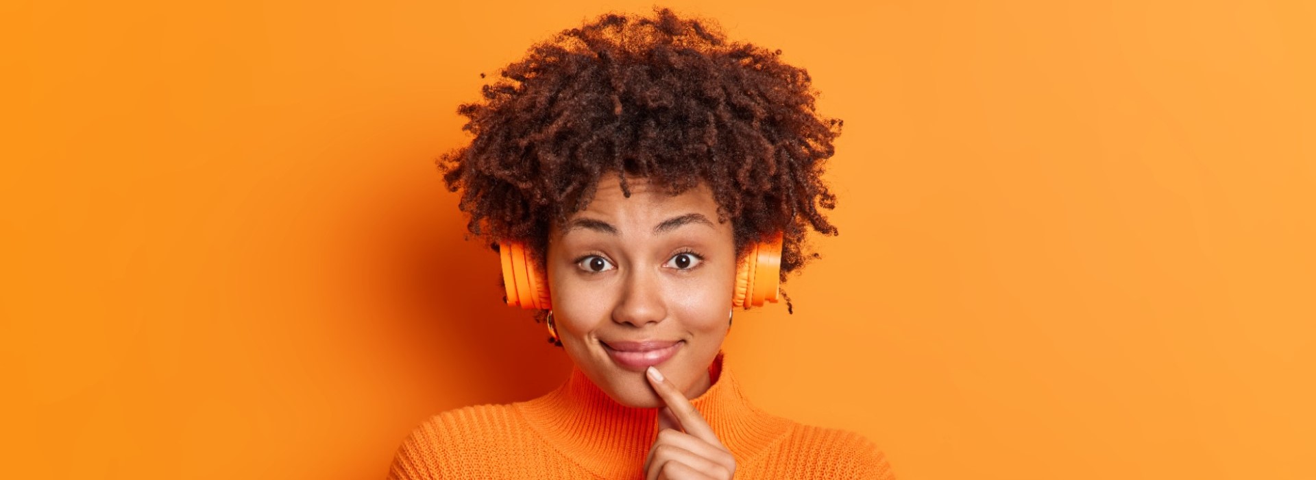 Imagem com fundo laranja. À frente uma mulher com fones de ouvido laranja e camiseta laranja leva o dedo indicador esquerdo à boca enquanto sorri. Fim da descrição.