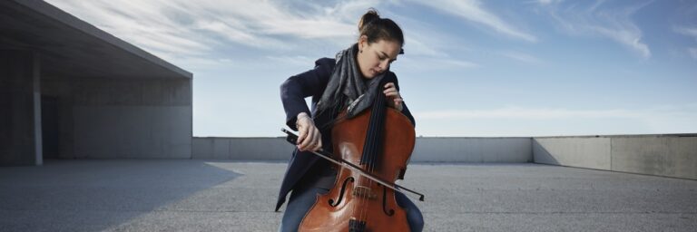 Mulher sentada tocando violoncelo. Ao fundo, um céu azul ensolorado com algumas nuvens, um chão de cimento atrás da mulher. Fim da descrição.