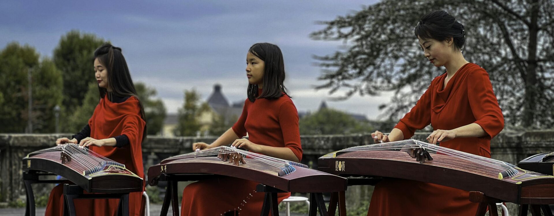 Three women in red dresses play guzheng instruments in an outdoor setting, with a serene blue sky backdrop.
